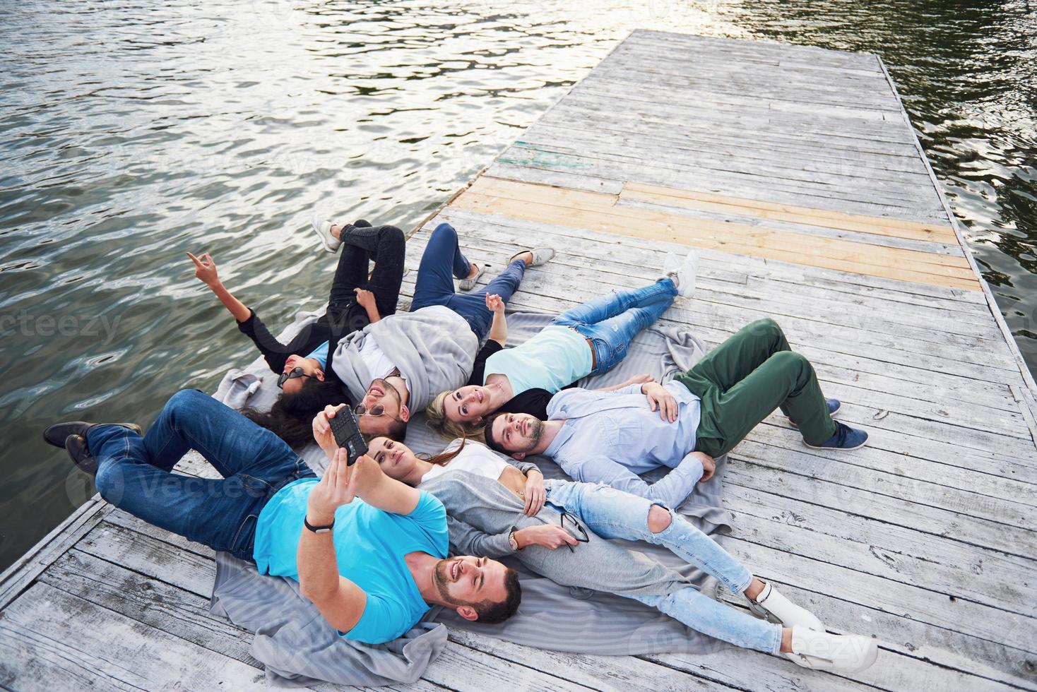 Gruppe schöner junger Leute, die Selfies machen, die auf dem Pier liegen, die besten Freunde von Mädchen und Jungen mit Vergnügen Konzept schafft emotionales Leben der Menschen. foto