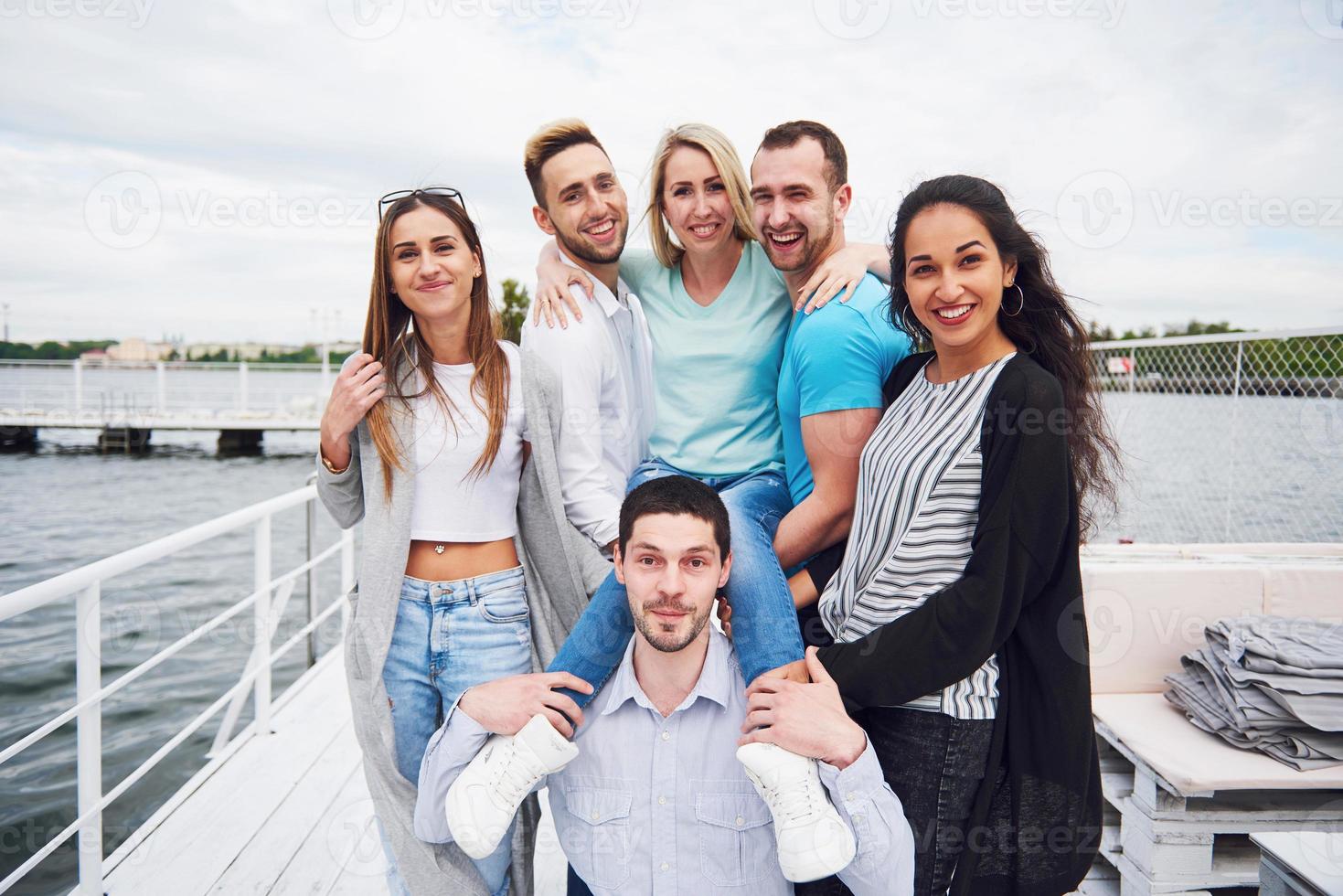 Porträt einer Gruppe junger Menschen, die am Rand der Seebrücke sitzen, draußen in der Natur. Freunde, die ein Spiel auf dem See genießen. foto