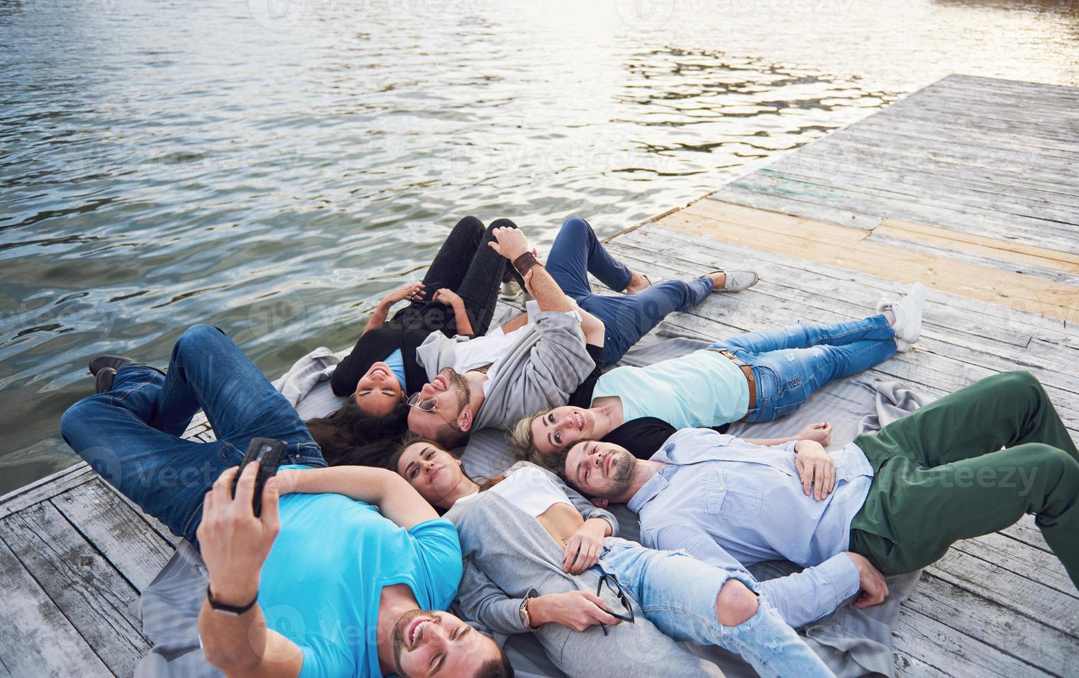 Porträt einer Gruppe junger Menschen, die am Rand der Seebrücke sitzen, draußen in der Natur. Freunde, die ein Spiel auf dem See genießen. foto