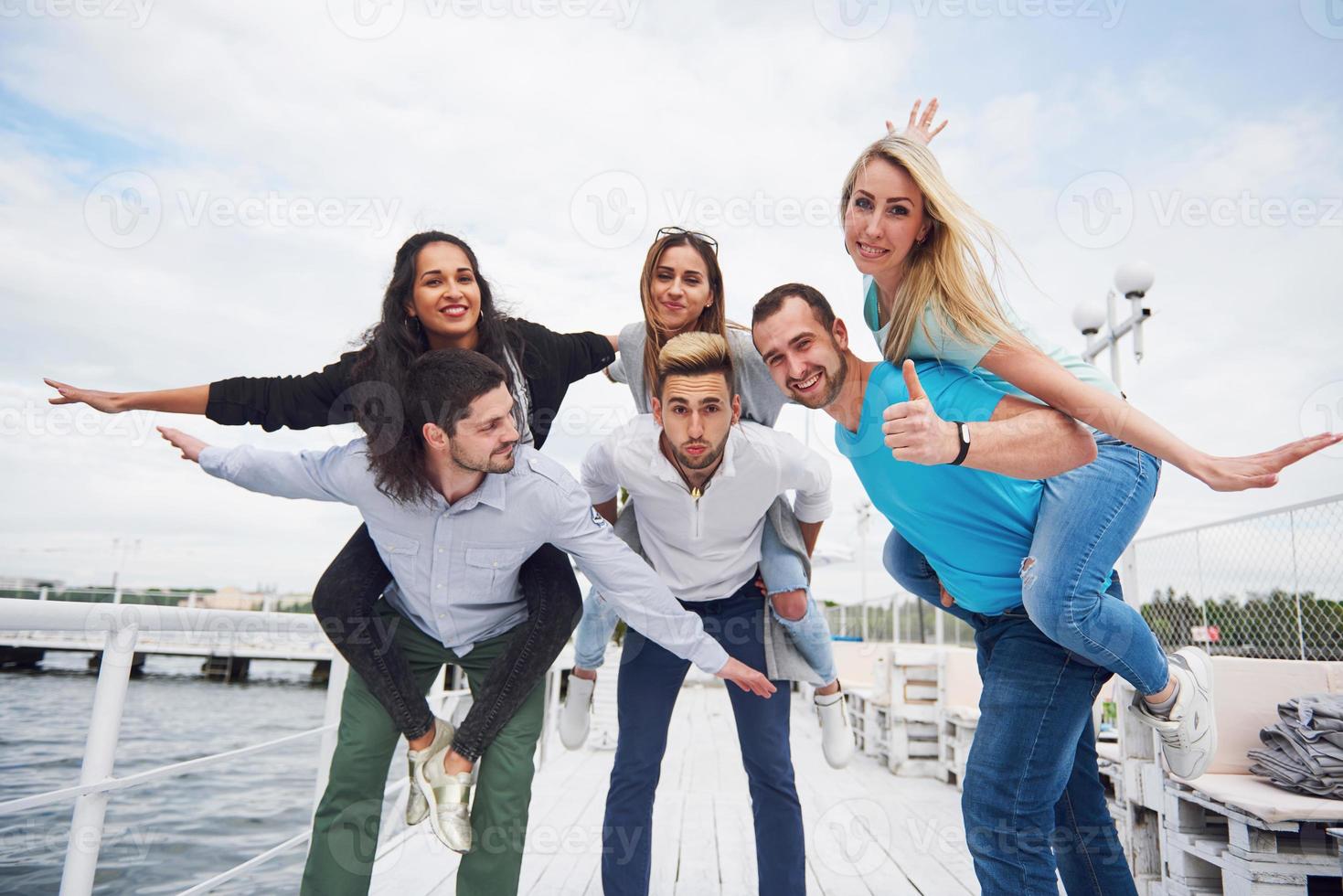 Porträt einer Gruppe junger Menschen, die am Rand der Seebrücke sitzen, draußen in der Natur. Freunde, die ein Spiel auf dem See genießen. foto