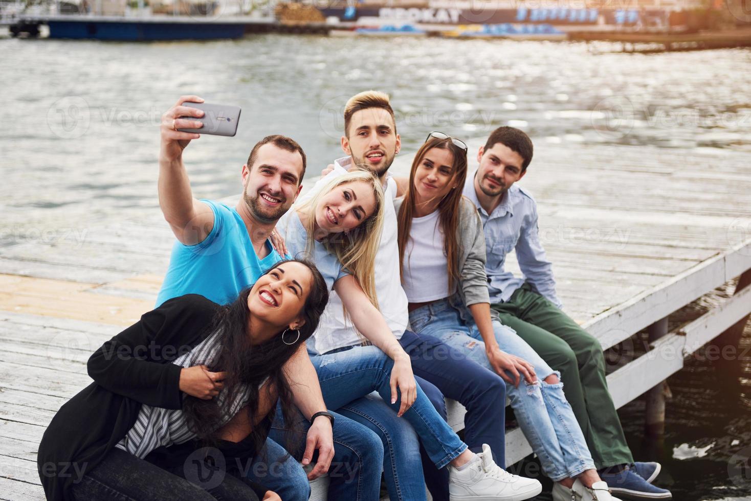 Eine Gruppe junger Leute sitzt am Rand des Piers und macht Selfies. Freunde, die ein Spiel auf dem See genießen foto