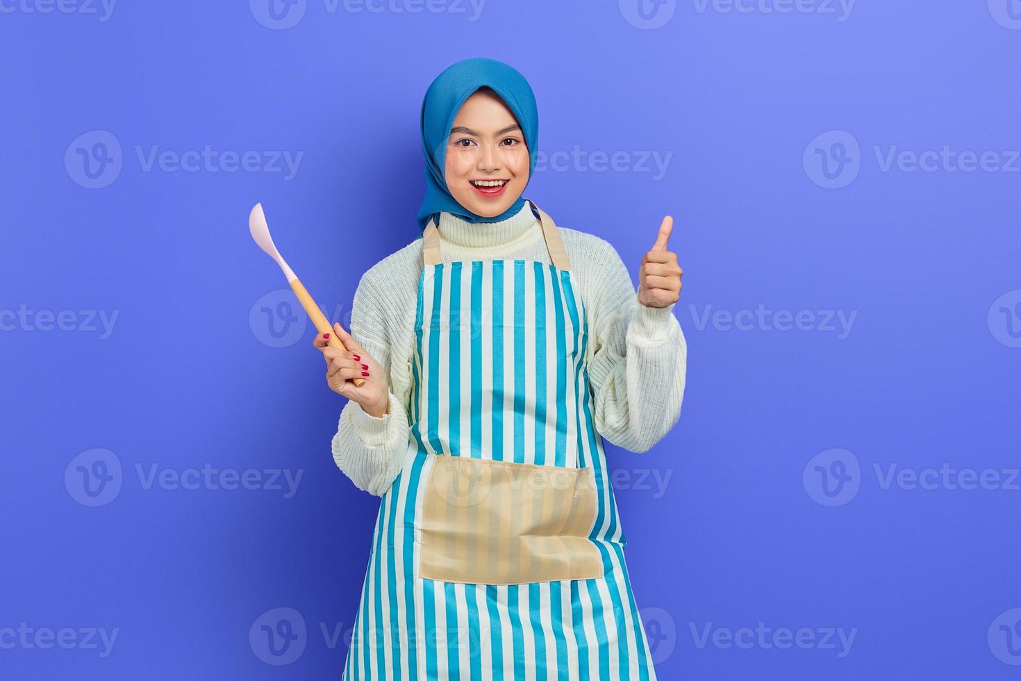 Fröhliche schöne asiatische Frau in weißem Pullover mit Schürze und Hijab, die einen Löffel hält und den Daumen isoliert über violettem Hintergrund zeigt. Housekeeping-Konzept foto