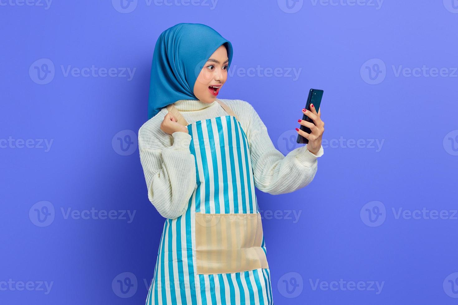 porträt einer aufgeregten jungen asiatischen muslimischen frau in den 20er jahren, die hijab und schürze trägt und ein handy mit einer siegergeste hält, die isoliert auf violettem hintergrund gemacht wird. Menschen Hausfrau muslimischen Lifestyle-Konzept foto
