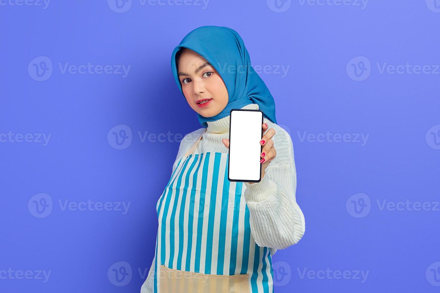 Porträt einer lächelnden jungen asiatischen muslimischen Frau mit Hijab und Schürze, die ein Handy mit leerem Bildschirm isoliert auf violettem Hintergrund hält. Menschen Hausfrau muslimischen Lifestyle-Konzept foto
