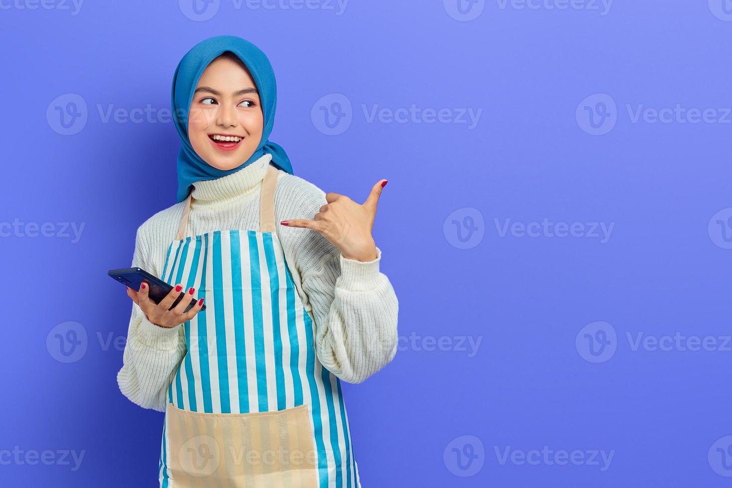 lächelnde junge asiatische muslimische frau, die hijab und schürze trägt, handy hält und telefongeste macht, wie sagt, ruf mich zurück, isoliert auf lila hintergrund. Menschen Hausfrau muslimischen Lifestyle-Konzept foto