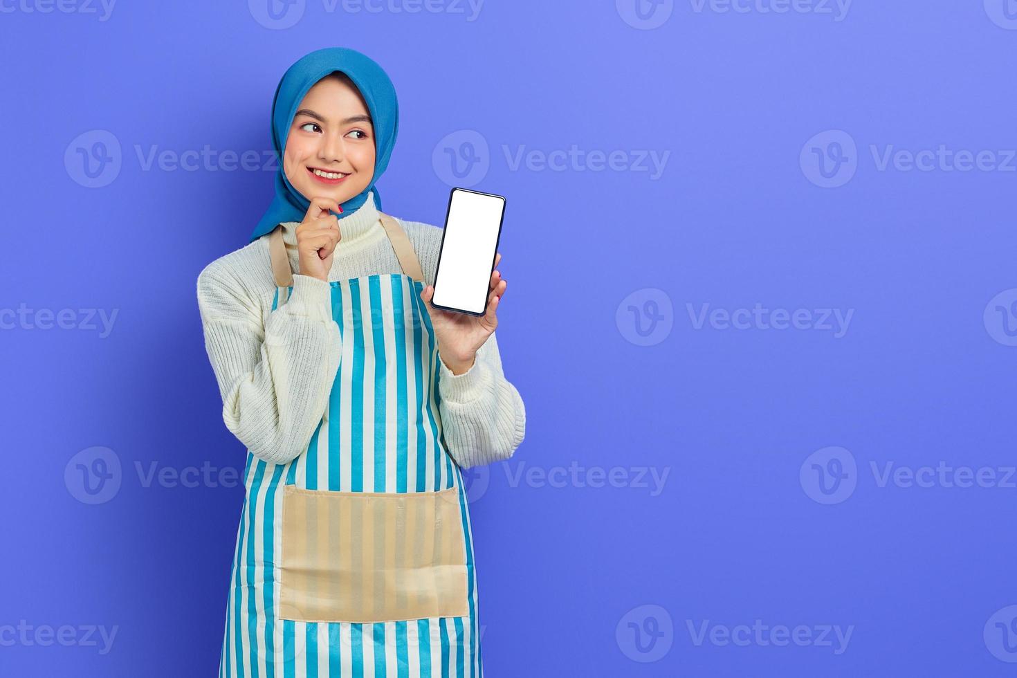 lächelnde junge asiatische muslimische Frau mit Hijab und Schürze, die ein Handy mit leerem Bildschirm hält, während sie das Kinn berührt und isoliert auf violettem Hintergrund zur Seite schaut. Menschen Hausfrau muslimischen Lifestyle-Konzept foto