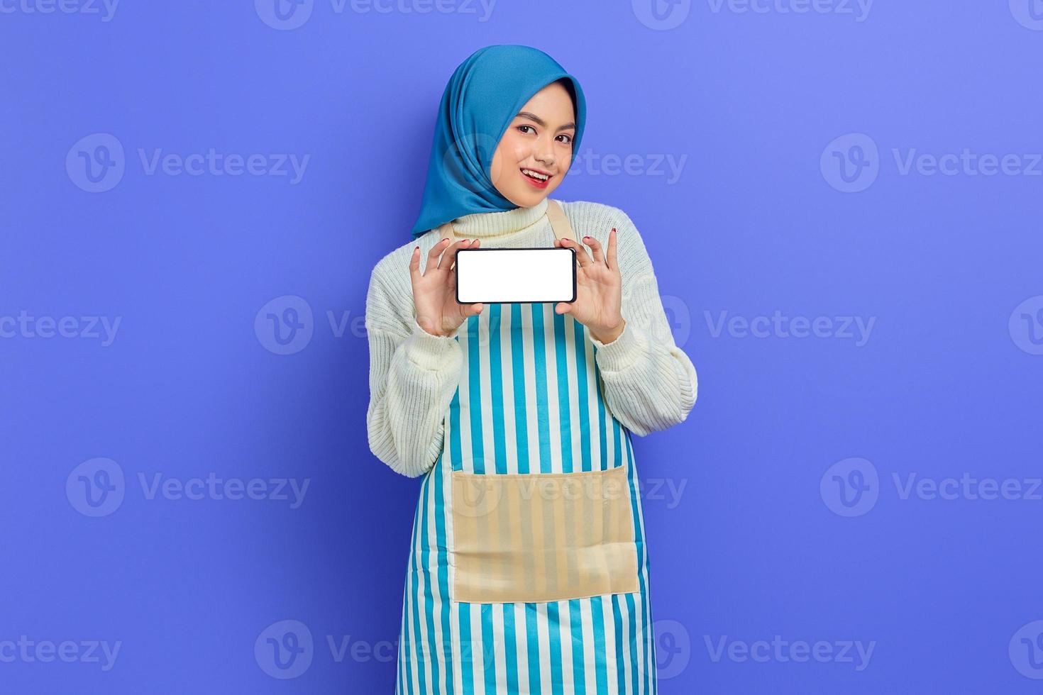 Fröhliche junge asiatische muslimische Frau mit Hijab und Schürze, die ein Handy mit leerem Bildschirm mit der Hand isoliert auf violettem Hintergrund hält. Menschen Hausfrau muslimischen Lifestyle-Konzept foto