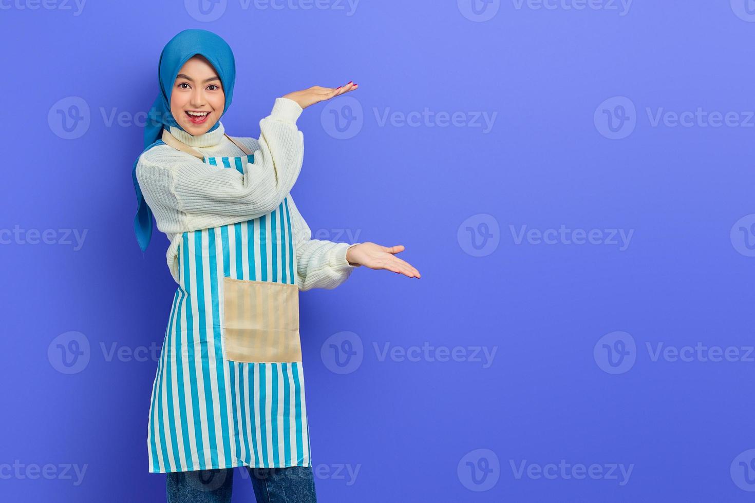 Porträt einer fröhlichen Hausfrau in Hijab und Schürze, die das Produkt mit der Hand zeigt und isoliert auf violettem Hintergrund in die Kamera blickt. Menschen Hausfrau muslimischen Lifestyle-Konzept foto