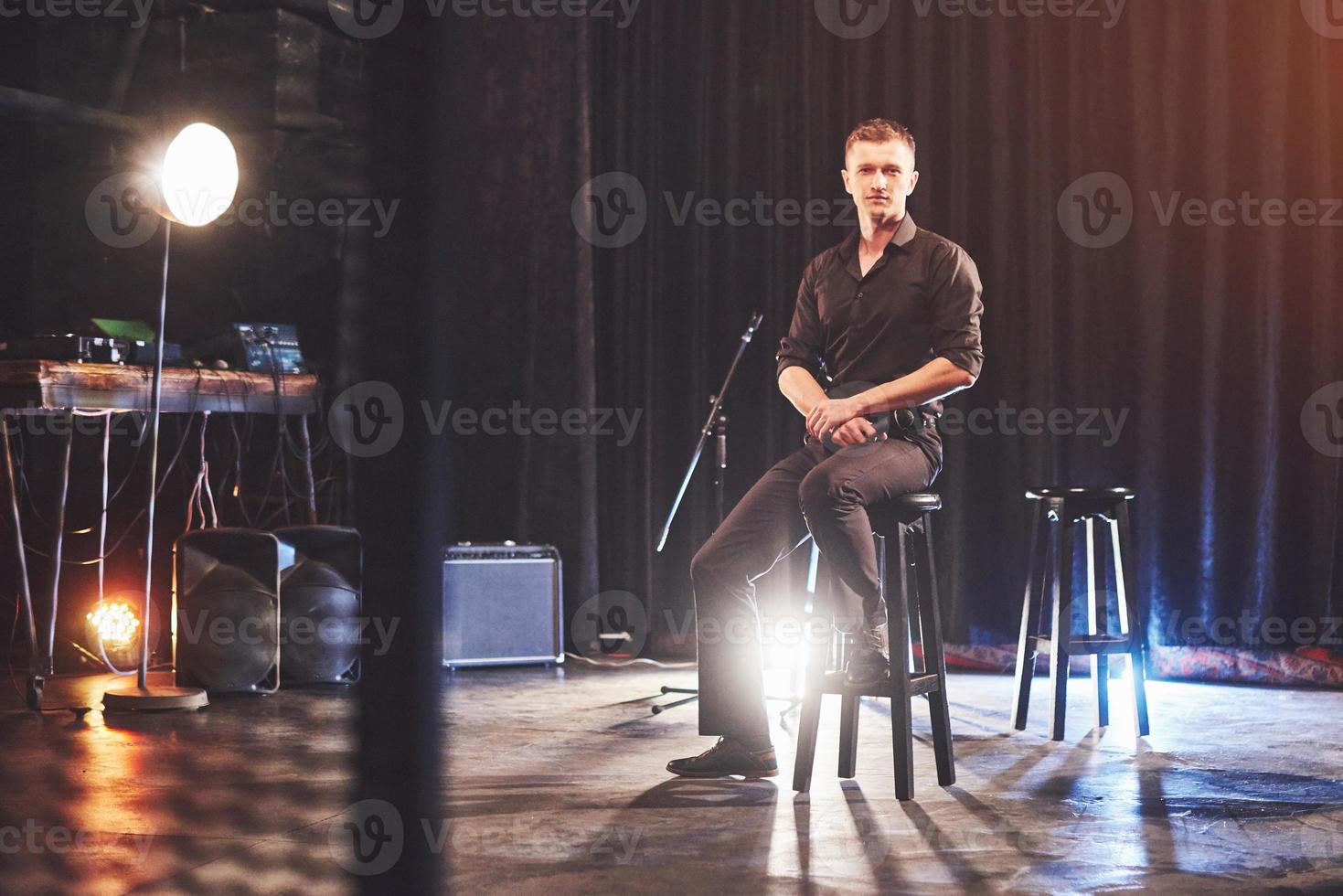 magischer Blick. hübscher junger Mann in schwarzer Kleidung, der in die Kamera blickt, während er auf einem Stuhl in der Nähe des dunklen Raums mit Licht sitzt foto