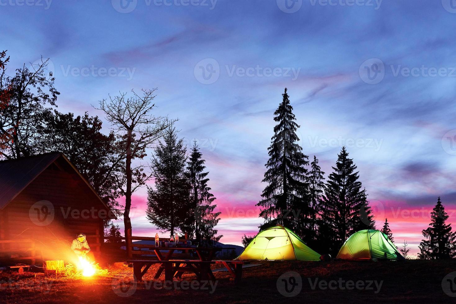 Nachtcamping. touristen ruhen sich an einem lagerfeuer in der nähe von beleuchtetem zelt und holzhaus unter einem erstaunlichen nachthimmel voller sterne und milchstraße aus foto