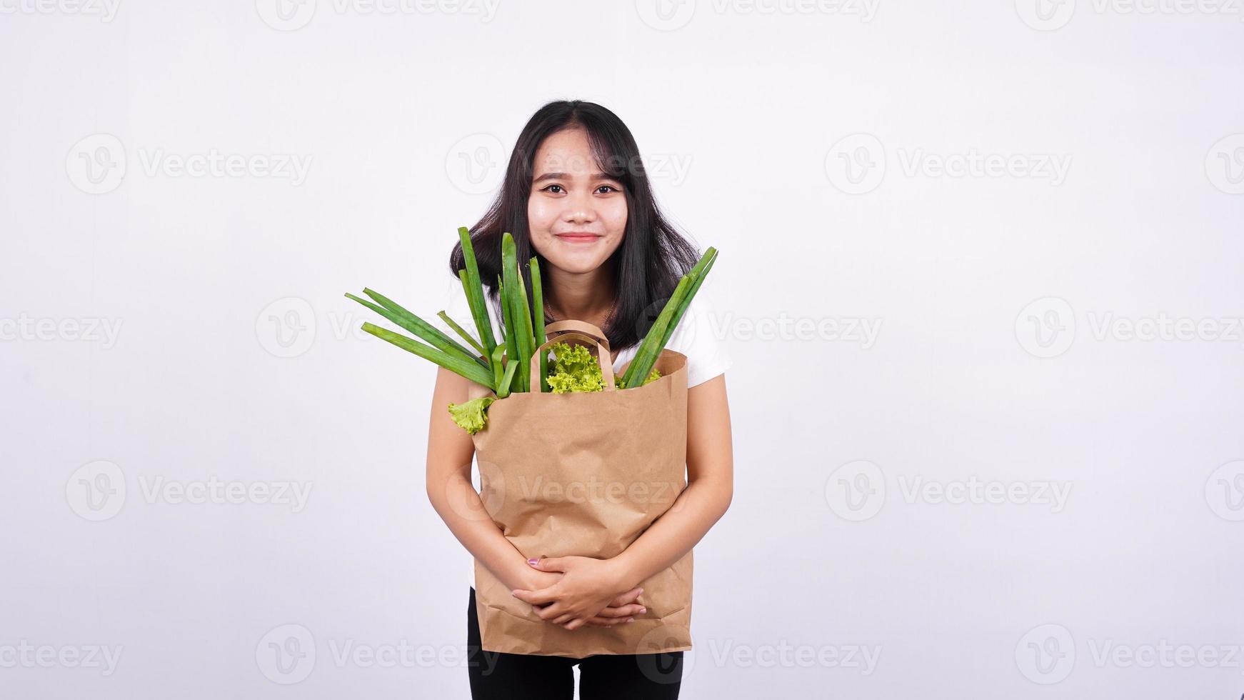 Schöne asiatische Frau, die mit Papiertüte mit frischem Gemüse mit isoliertem weißem Hintergrund lächelt foto