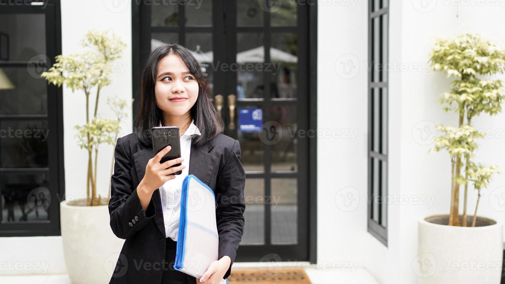 Schöne junge asiatische Geschäftsfrau im Anzug, die im Café mit Telefon und braunem Umschlag steht foto