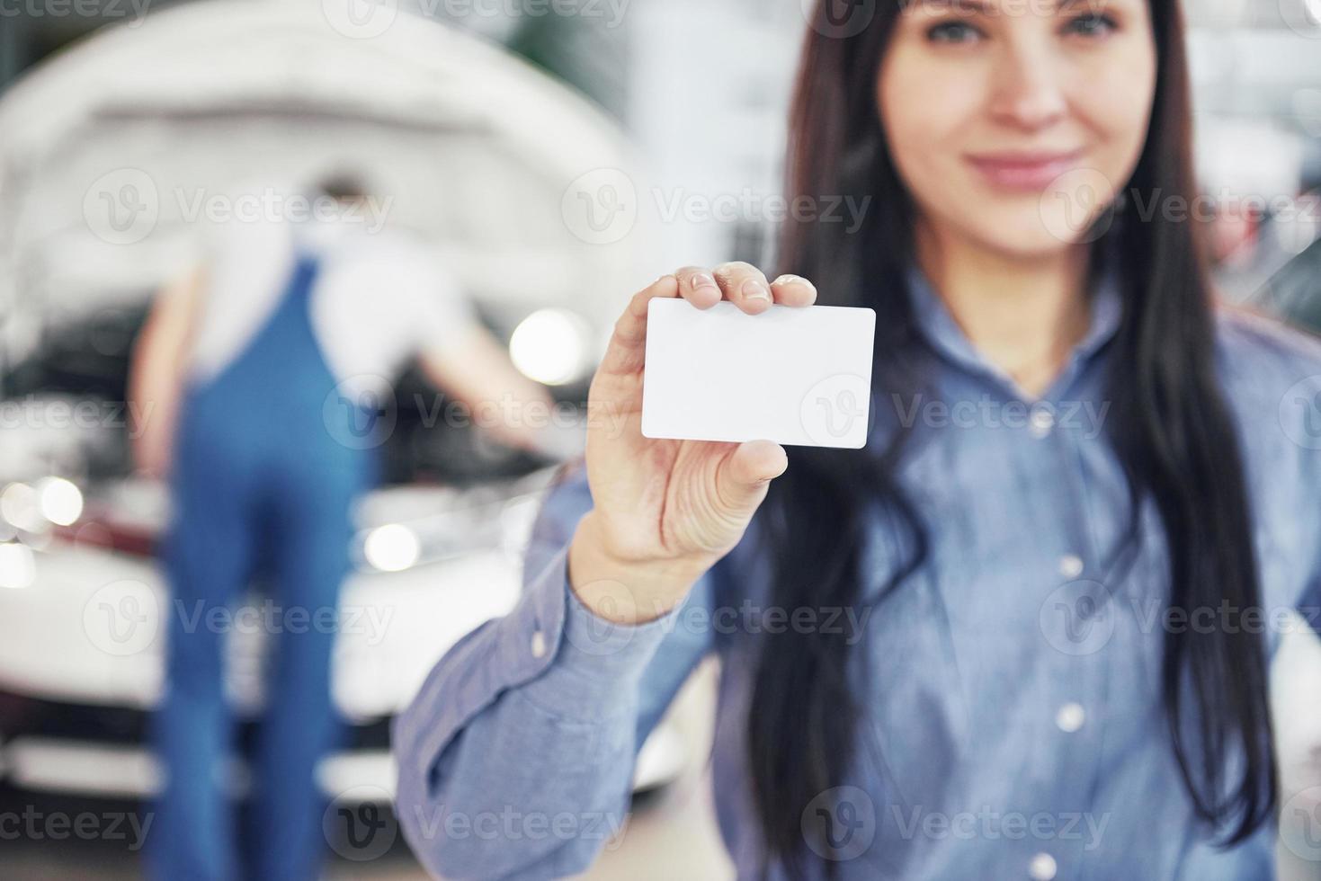 Eine schöne Frau hält eine Visitenkarte des Autoservice-Centers. der mechaniker inspiziert das auto im hintergrund unter der motorhaube foto