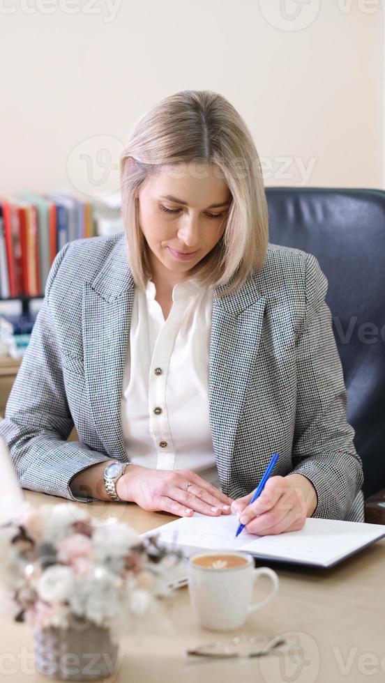 Linkshänderin schreibt in einem Notizbuch, das einen Arbeitstag plant. arbeitgeberin, die morgens formelle chothes im büro trägt. vertikaler Schuss foto