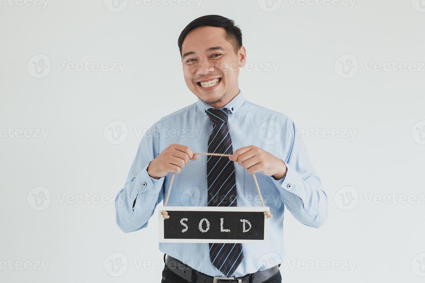 lächelnder Verkaufsmann, der blaues Hemd und Krawatte trägt, posiert mit dem Tragen von verkauftem Schild in die Kamera auf weißem Hintergrund foto