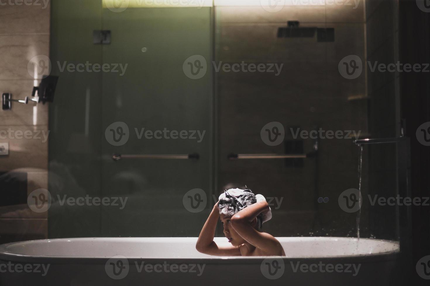 asiatischer junge, der auf der badewanne mit schaum auf seinem kopf shampooniert foto