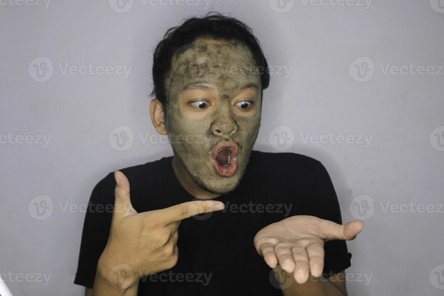 Lächelnder asiatischer Mann, der in die Hand zeigt, wenn er eine Schönheitsgesichtsmaske verwendet foto