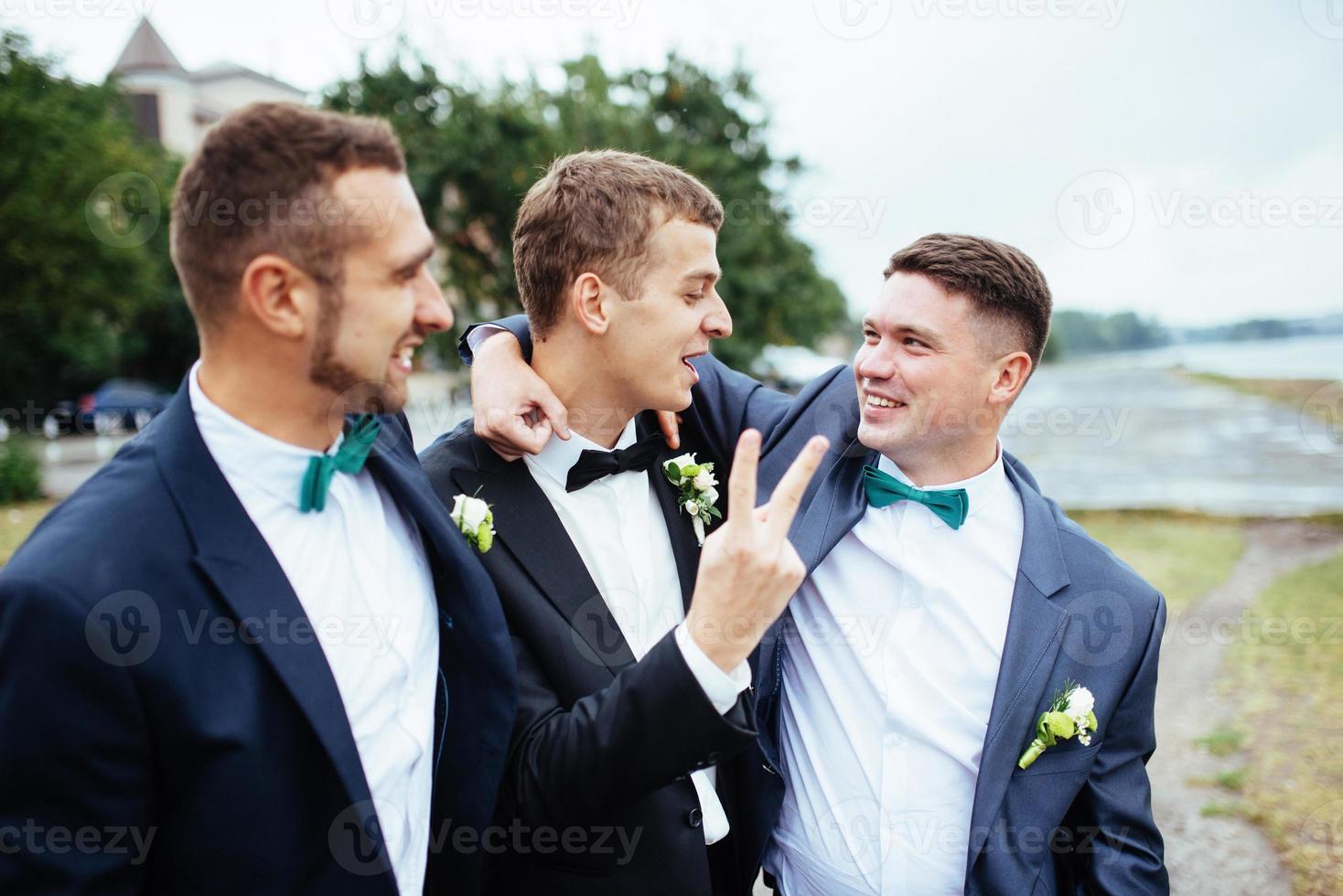 selbstbewusster, lächelnder, gutaussehender Bräutigam im schwarzen Anzug mit zwei Groomsma foto