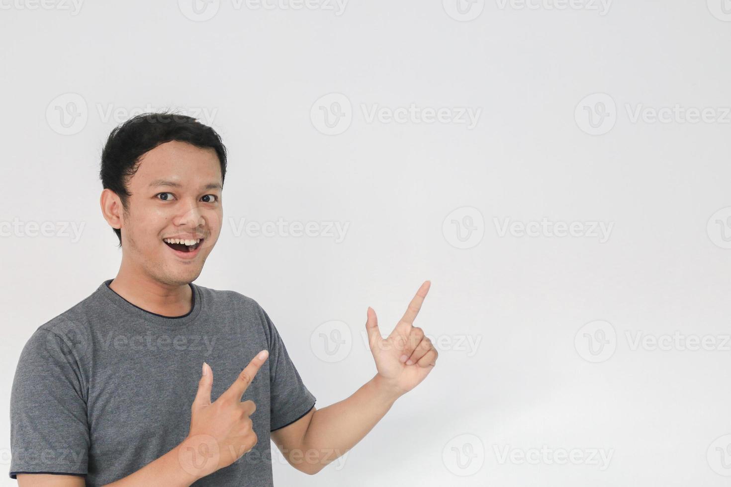 wow und überraschtes gesicht mit lächeln eines jungen asiatischen mannes im grauen t-shirt mit handzeig auf leeren raum. foto