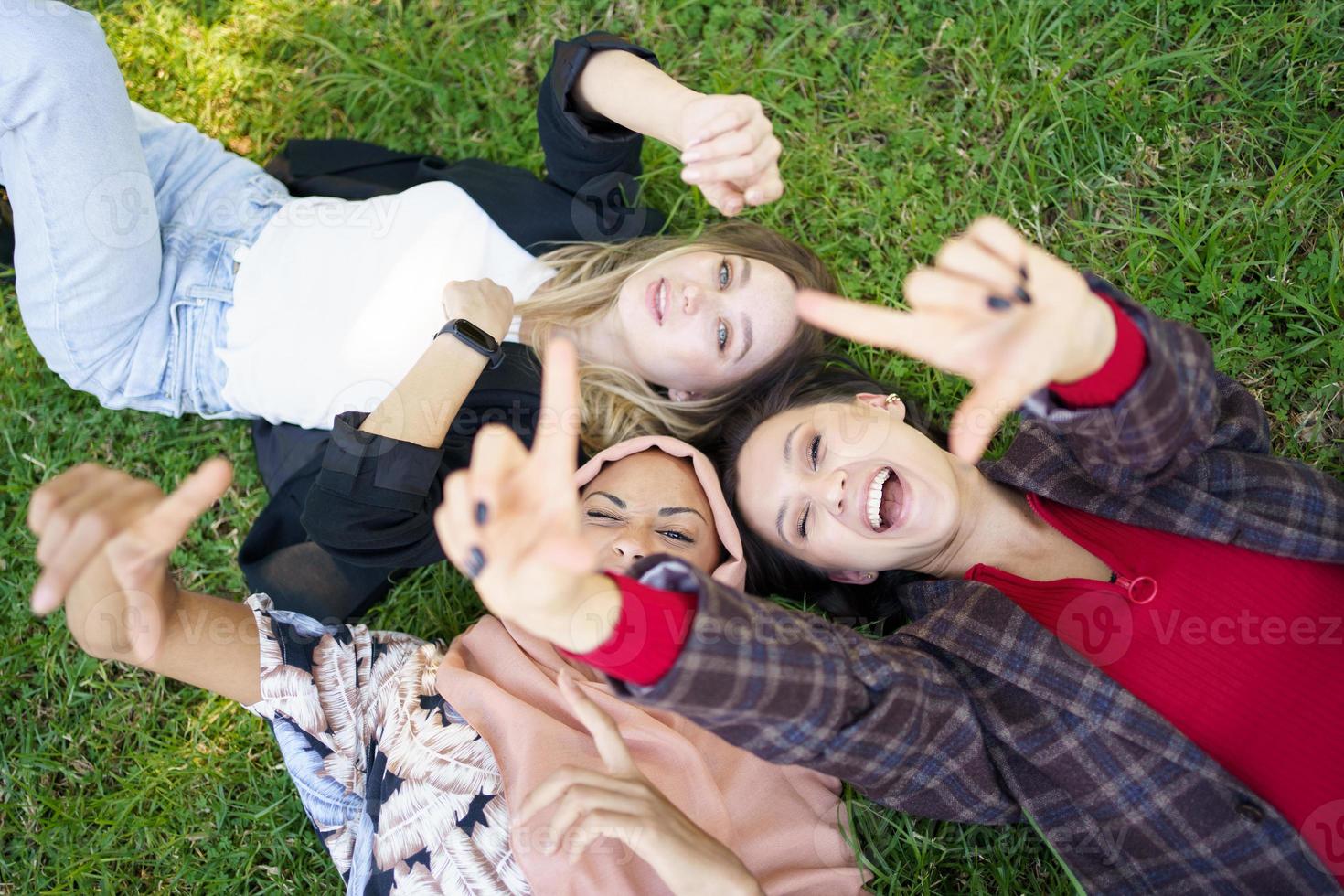 fröhliche damen, die kamera gestikulieren, während sie auf gras liegen foto