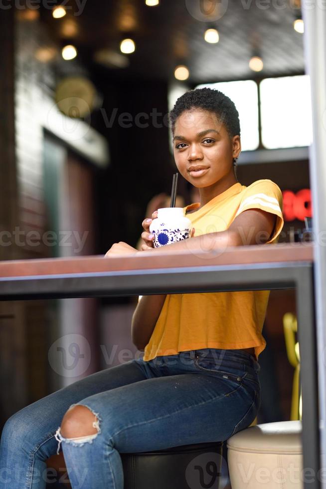 lächelnde schwarze Frau mit sehr kurzen Haaren, die einen Cocktail in einem städtischen Café trinkt. foto