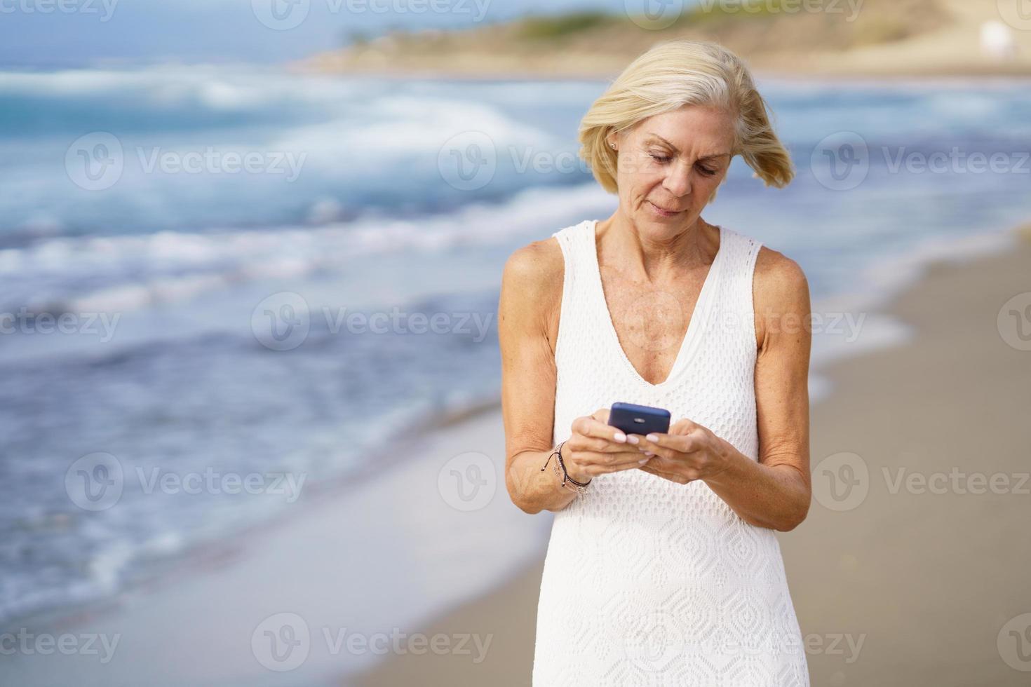 Seniorin, die mit einem Smartphone am Strand spazieren geht. foto