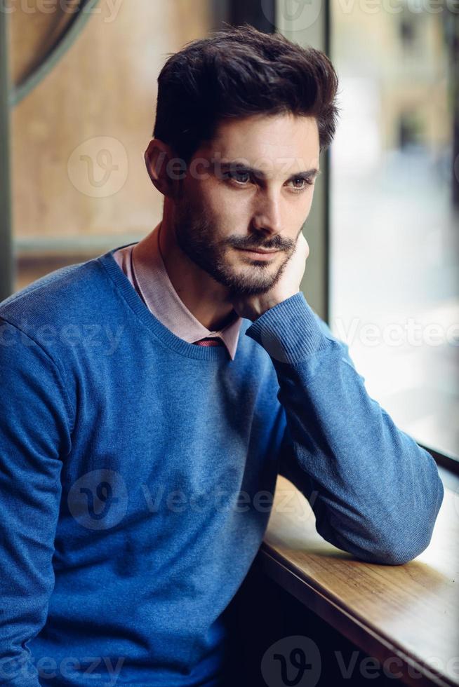 nachdenklicher mann mit blauem pullover mit verlorenem blick in der nähe eines fensters foto