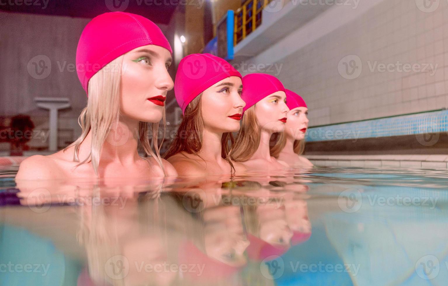 Vier wunderschöne, schlanke, junge, stilvolle Frauen in farbenfrohen Badeanzügen und rosa Schwimmhüten im Schwimmbad foto