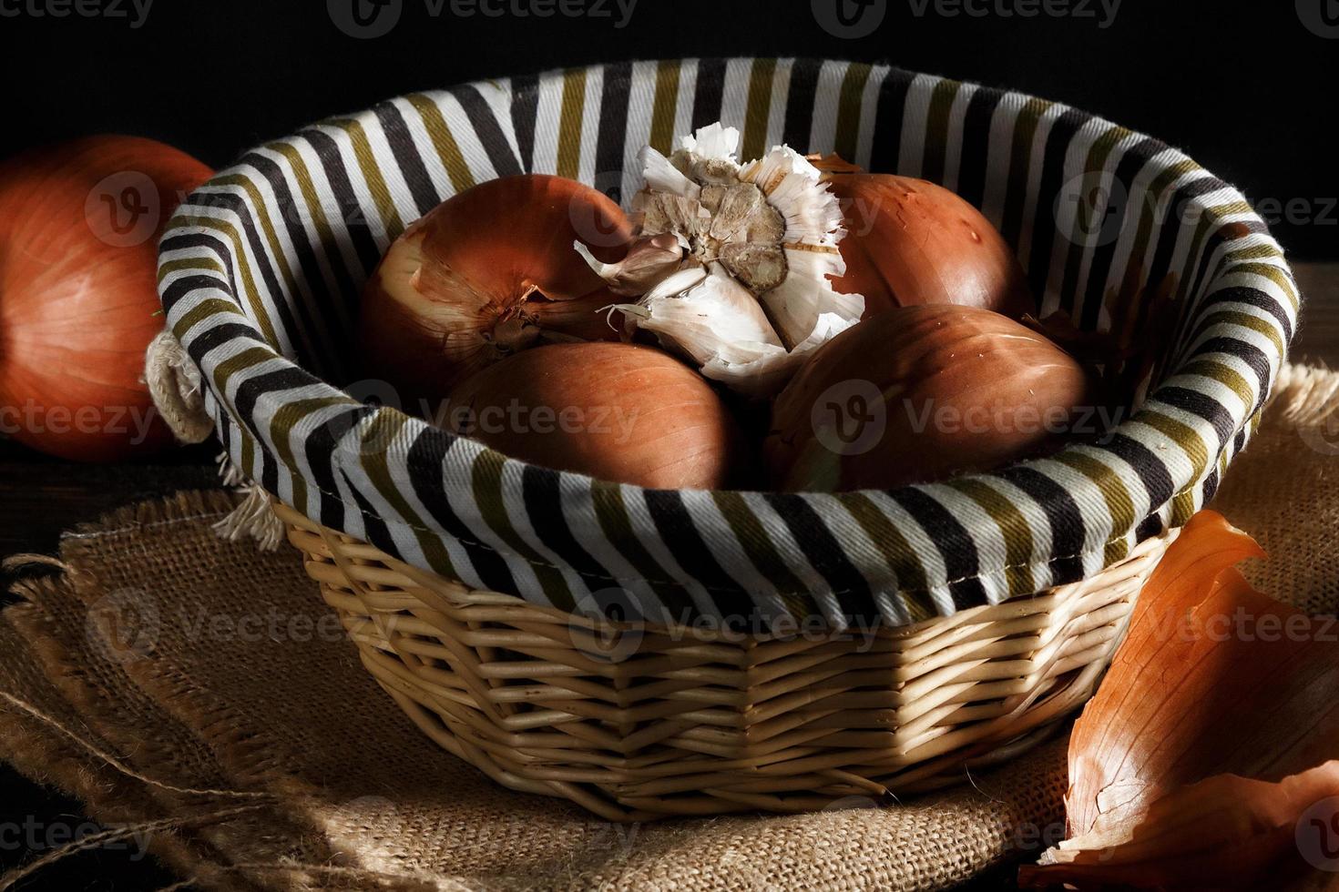 Zwiebeln und Knoblauch in einem Weidenkorb auf Holzbrettern und Sackleinen auf schwarzem Hintergrund. rustikaler Stil. horizontales Bild. foto