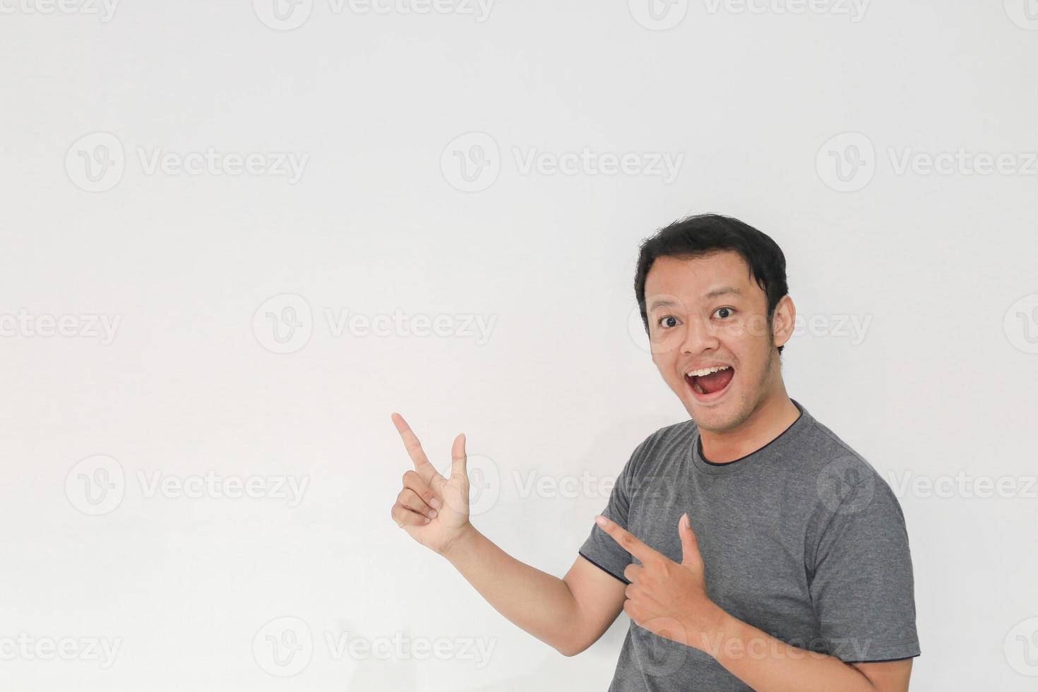 wow und überraschtes gesicht mit lächeln eines jungen asiatischen mannes im grauen t-shirt mit handzeig auf leeren raum. foto