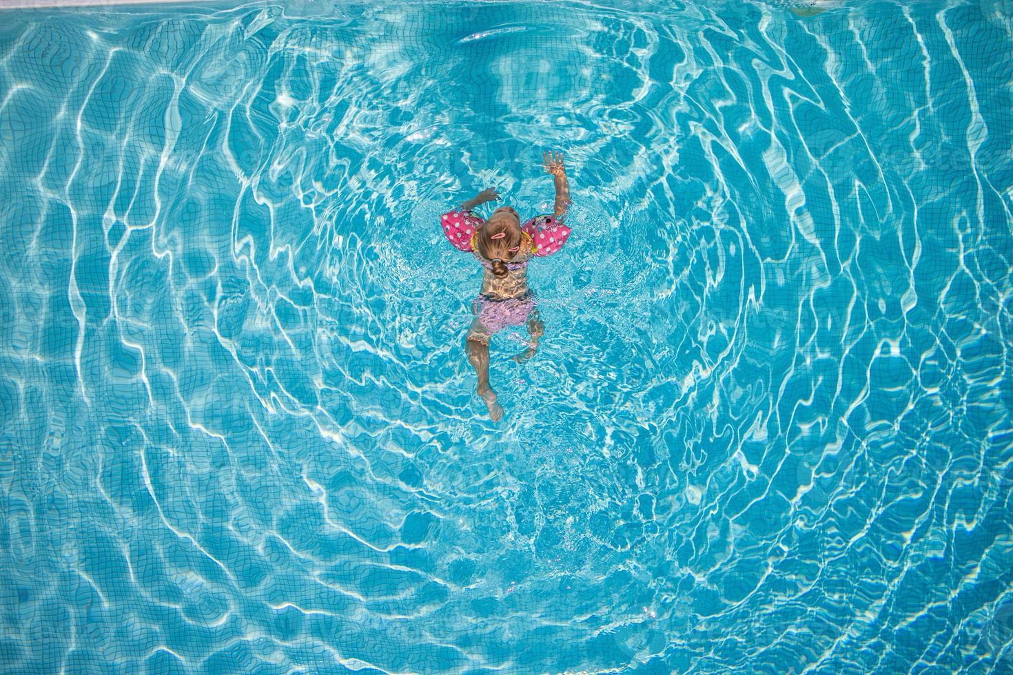 Süßes kleines Kleinkind, das im türkisfarbenen Pool mit aufblasbaren Armhilfen schwimmt, Draufsicht. foto