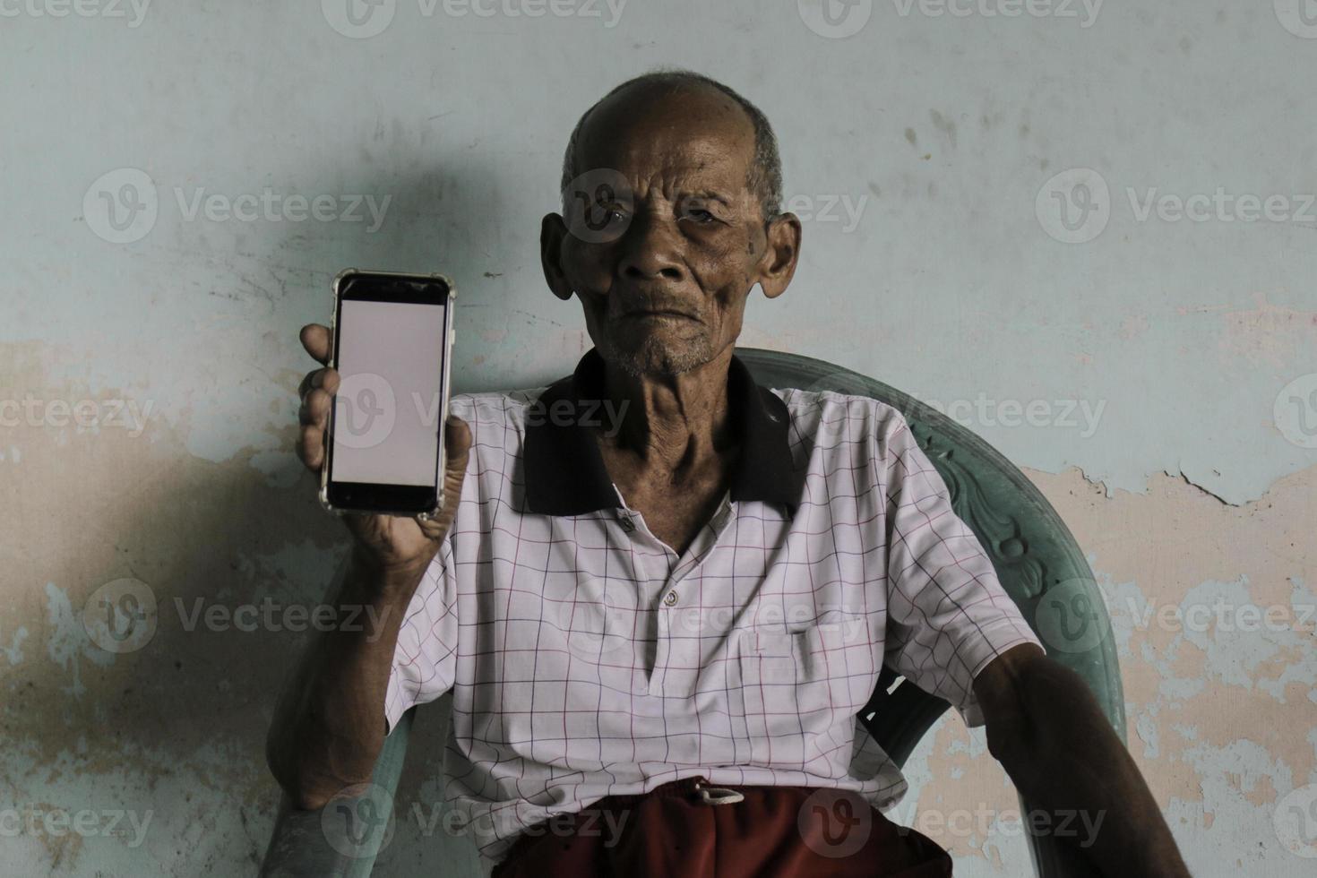 ein porträt des indonesischen alten opas, der ein smartphone mit weißem bildschirm zeigt. foto