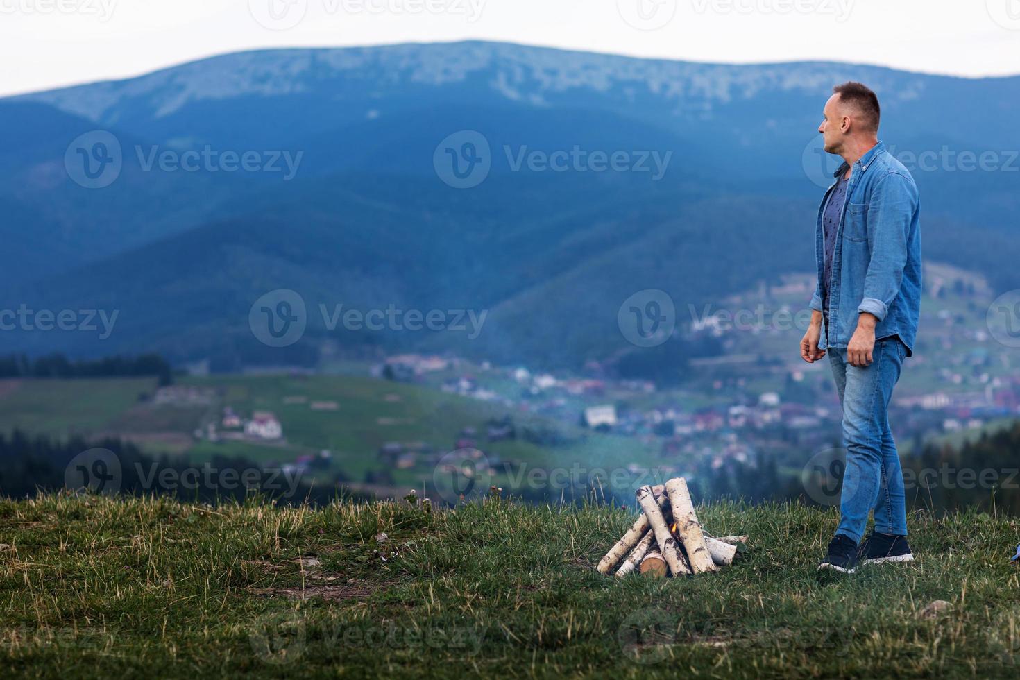 junger Mann, der an einem brennenden Feuer unter dem Abendhimmel in den Bergen steht. aktives Lifestyle-Konzept foto