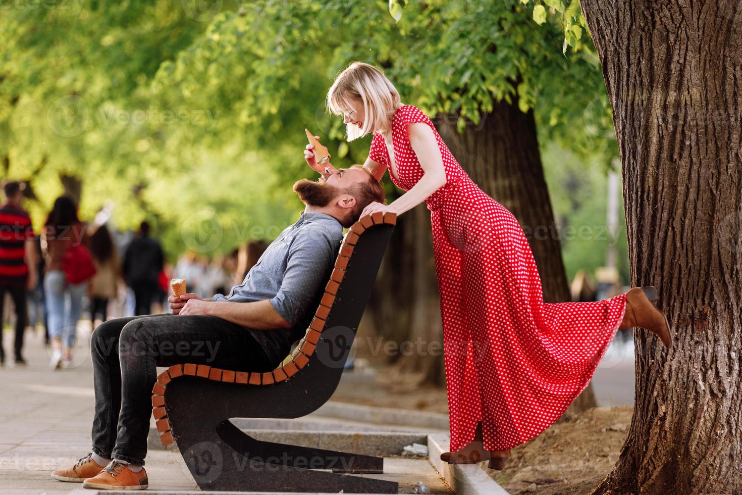 lächelnde Hipster-Paar, die Spaß hat und Eis in der Stadt isst. stylischer junger Mann mit Bart sitzt auf einer Holzbank und blonde Frau im roten Kleid albert herum und spielt mit ihm foto