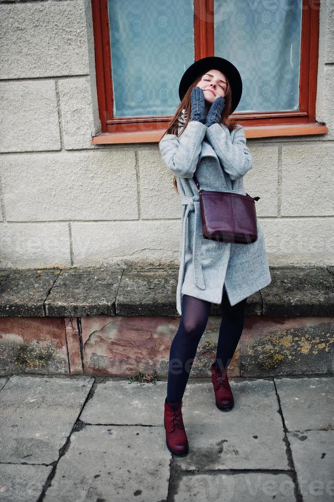 Junges Modelmädchen in grauem Mantel und schwarzem Hut mit Lederhandtasche auf den Schultern vor dem Fenster des alten Hauses in der Straße der Stadt. foto