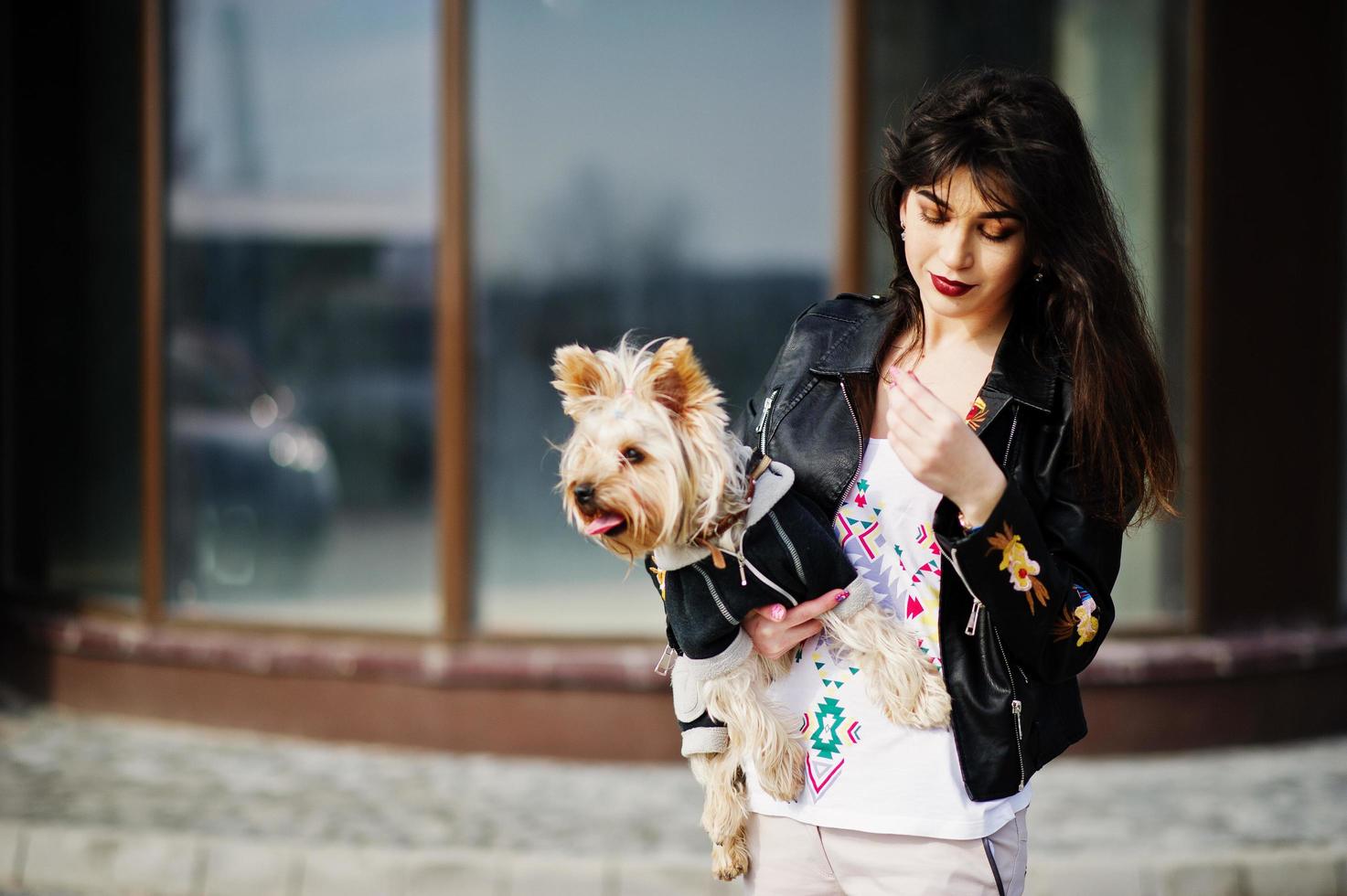 Brünette Zigeunerin mit Yorkshire-Terrier-Hund posierte gegen Haus mit großen Fenstern. Model Wear auf Lederjacke und T-Shirt mit Ornament, Hose. foto