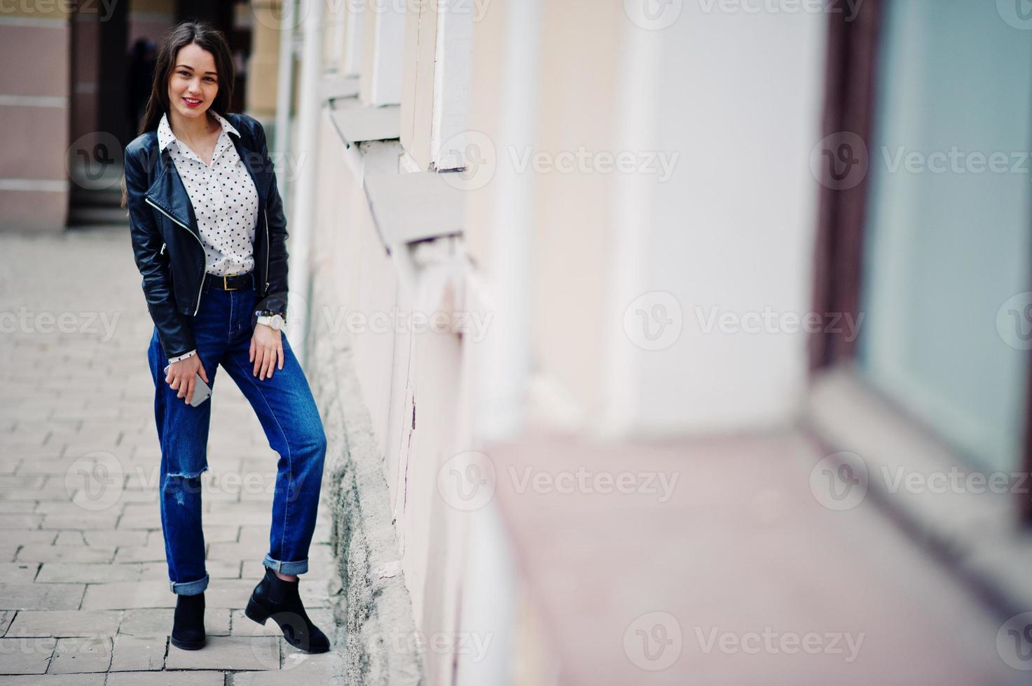 Porträt stilvoller Kleidung junger Mädchen auf Lederjacke und zerrissenen Jeans mit Handy zur Hand. Straßenmode-Model-Stil. foto