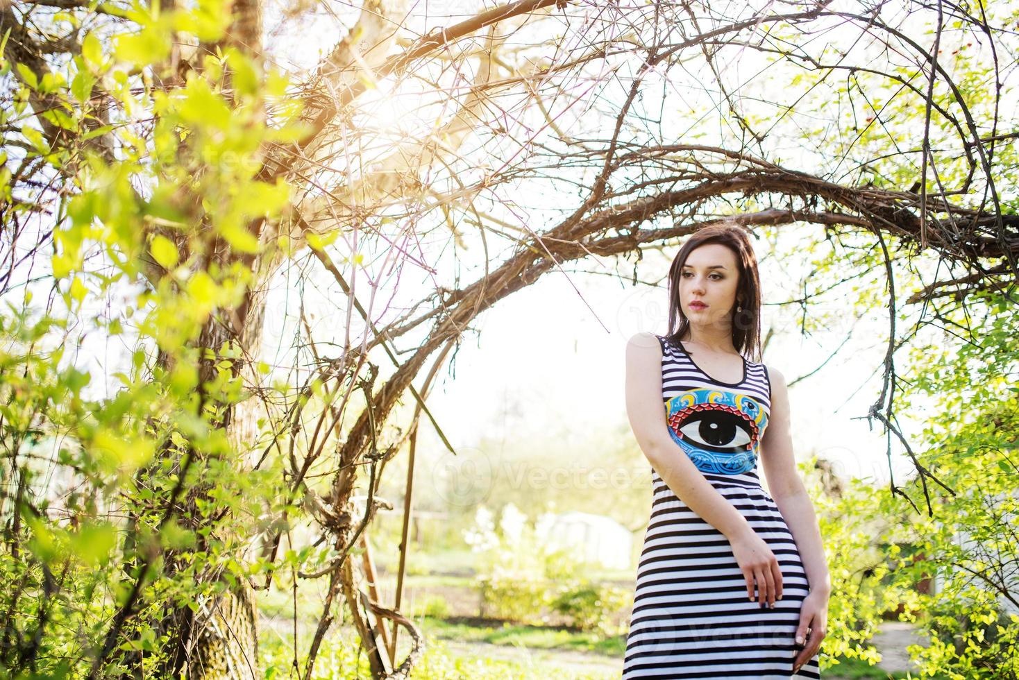 brünettes vorbildliches mädchen im kleid mit streifen posierte in der nähe des bogens vom baum. foto