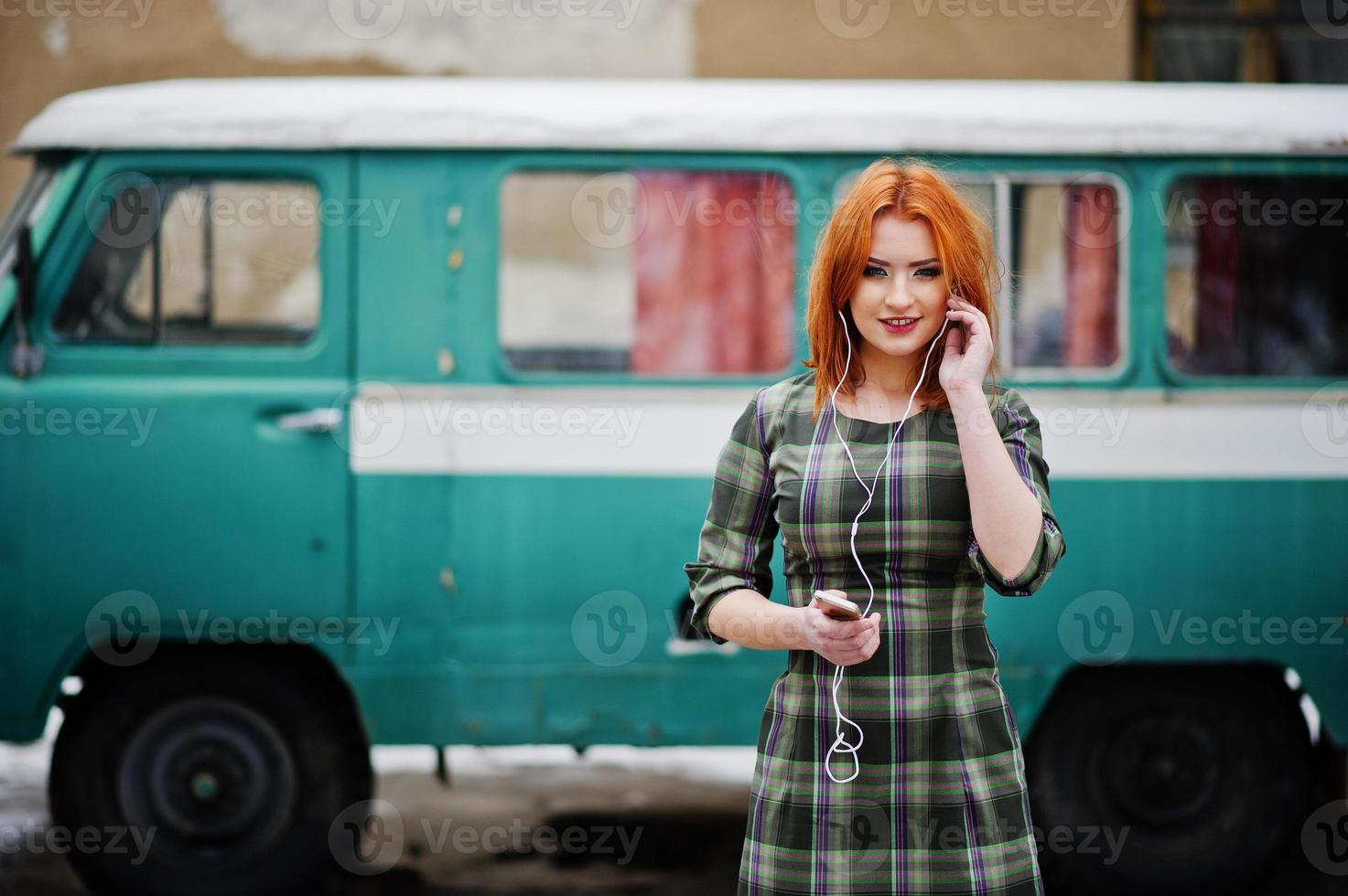 junge rothaarige Mädchendame mit Handy und Kopfhörern, die auf kariertem Kleid Hintergrund trägt alter Vintage-Türkis-Minivan-Bus. foto