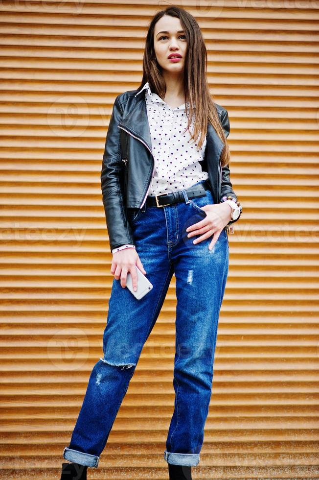 Porträt von stilvollen jungen Mädchen auf Lederjacke und zerrissenen Jeans mit Handy zur Hand Hintergrund-Shutter-Textur. Straßenmode-Model-Stil. foto