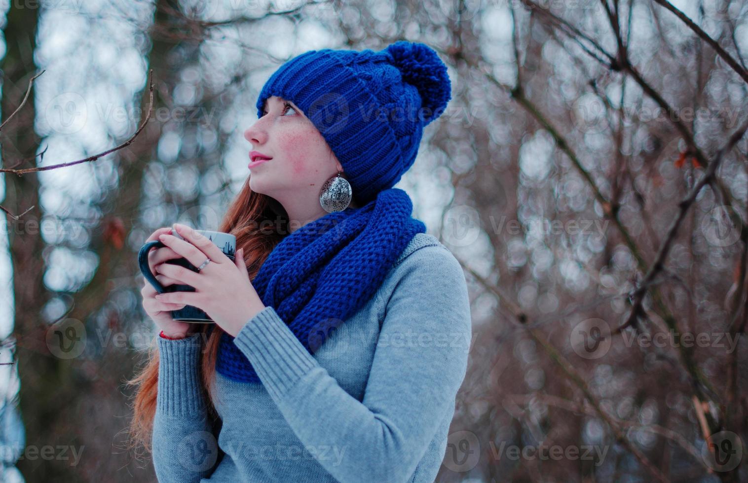 Porträt eines jungen Mädchens mit roten Haaren und Sommersprossen, das am Wintertag eine blau gestrickte Wollmütze und einen Schal mit einer Tasse Tee trägt. foto