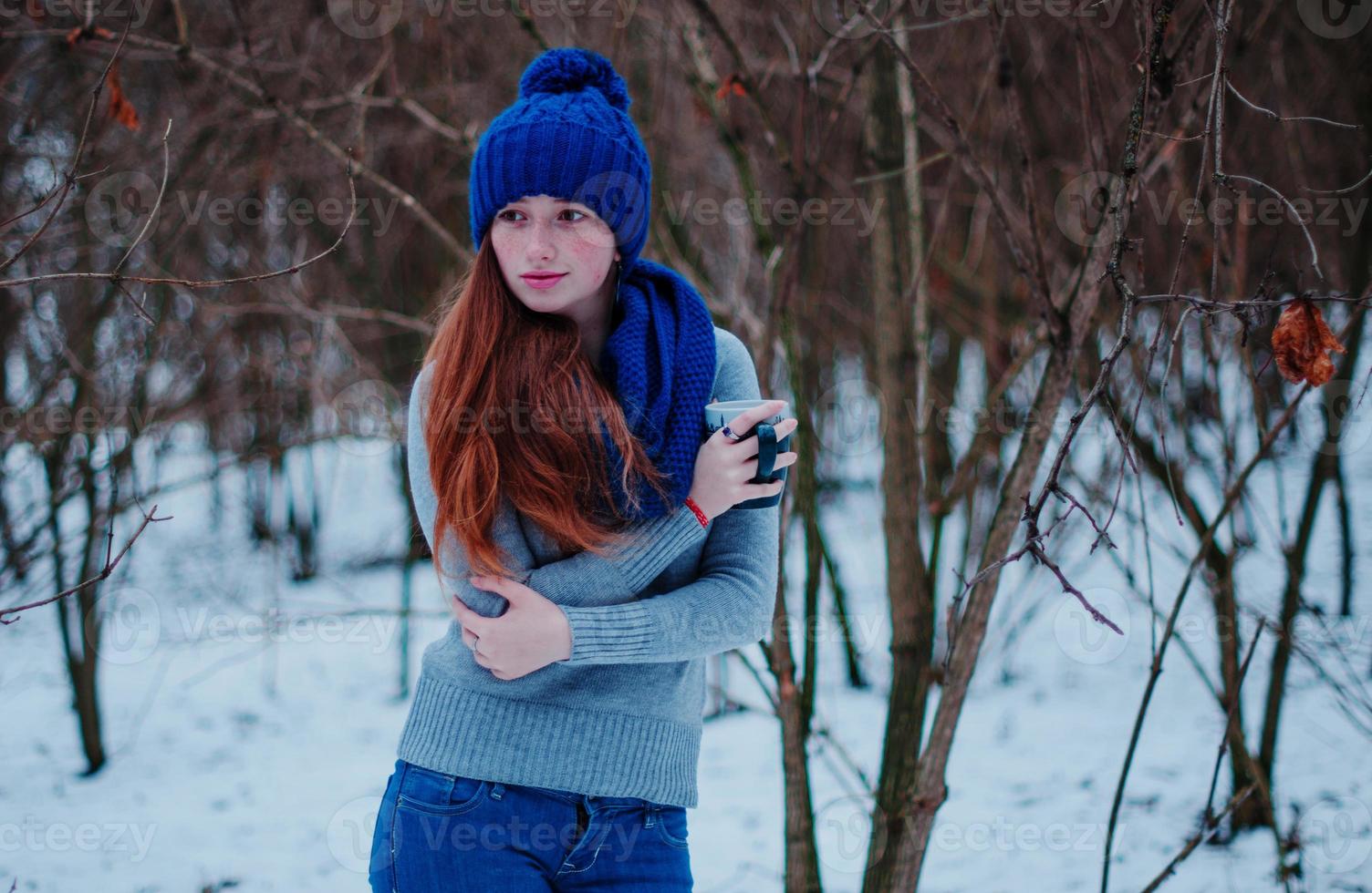 Porträt eines jungen Mädchens mit roten Haaren und Sommersprossen, das am Wintertag eine blau gestrickte Wollmütze und einen Schal mit einer Tasse Tee trägt. foto