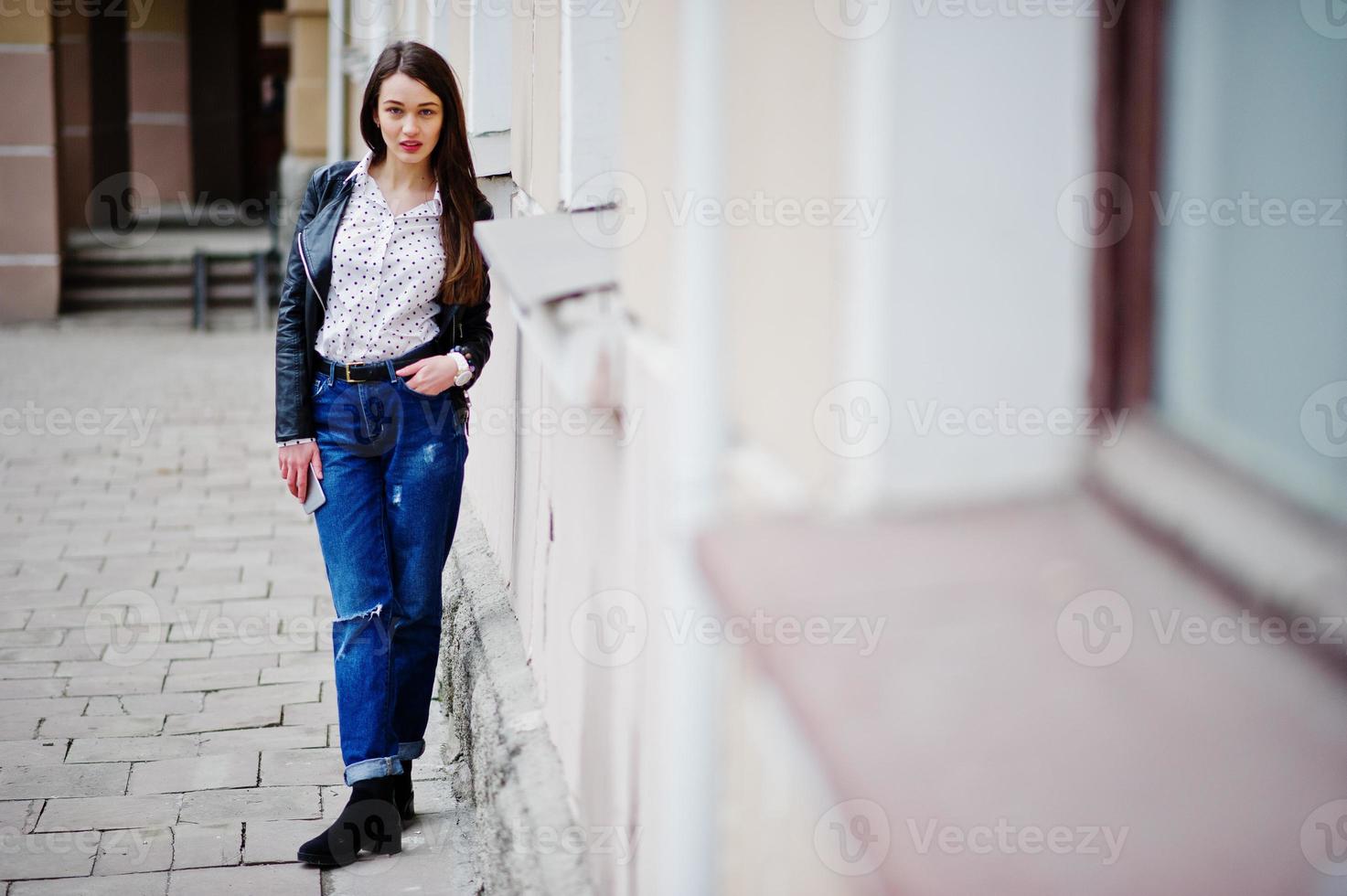 Porträt stilvoller Kleidung junger Mädchen auf Lederjacke und zerrissenen Jeans mit Handy zur Hand. Straßenmode-Model-Stil. foto