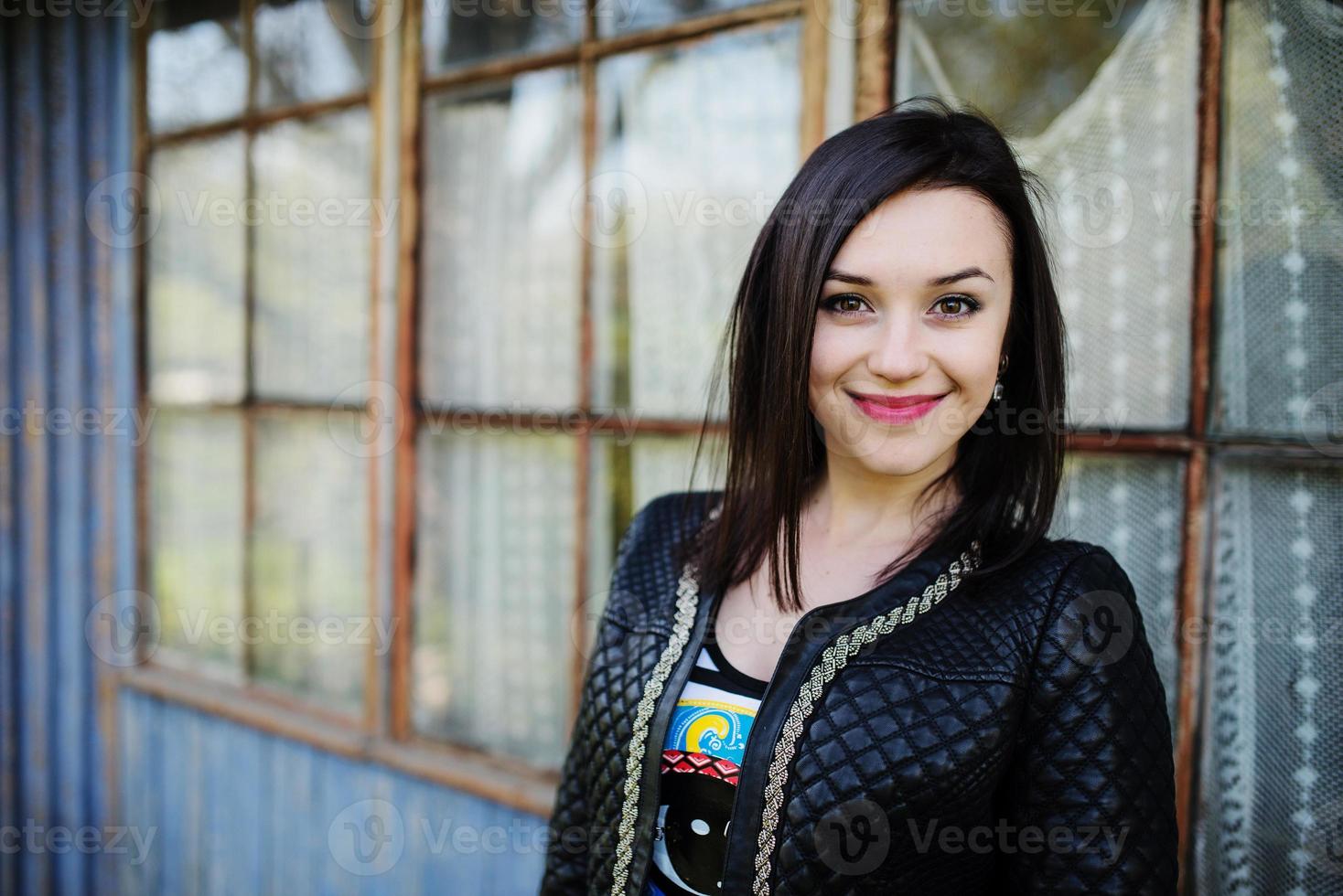 Brünette Modell Mädchen Lederjacke gestellt Hintergrund altes Holzhaus mit Fenstern. foto