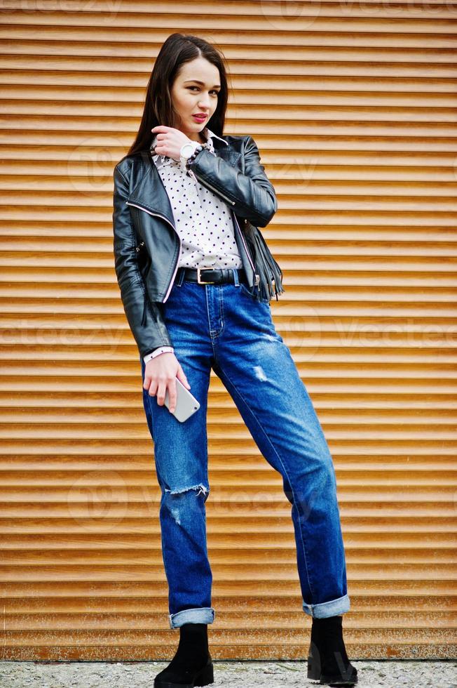 Porträt von stilvollen jungen Mädchen auf Lederjacke und zerrissenen Jeans mit Handy zur Hand Hintergrund-Shutter-Textur. Straßenmode-Model-Stil. foto