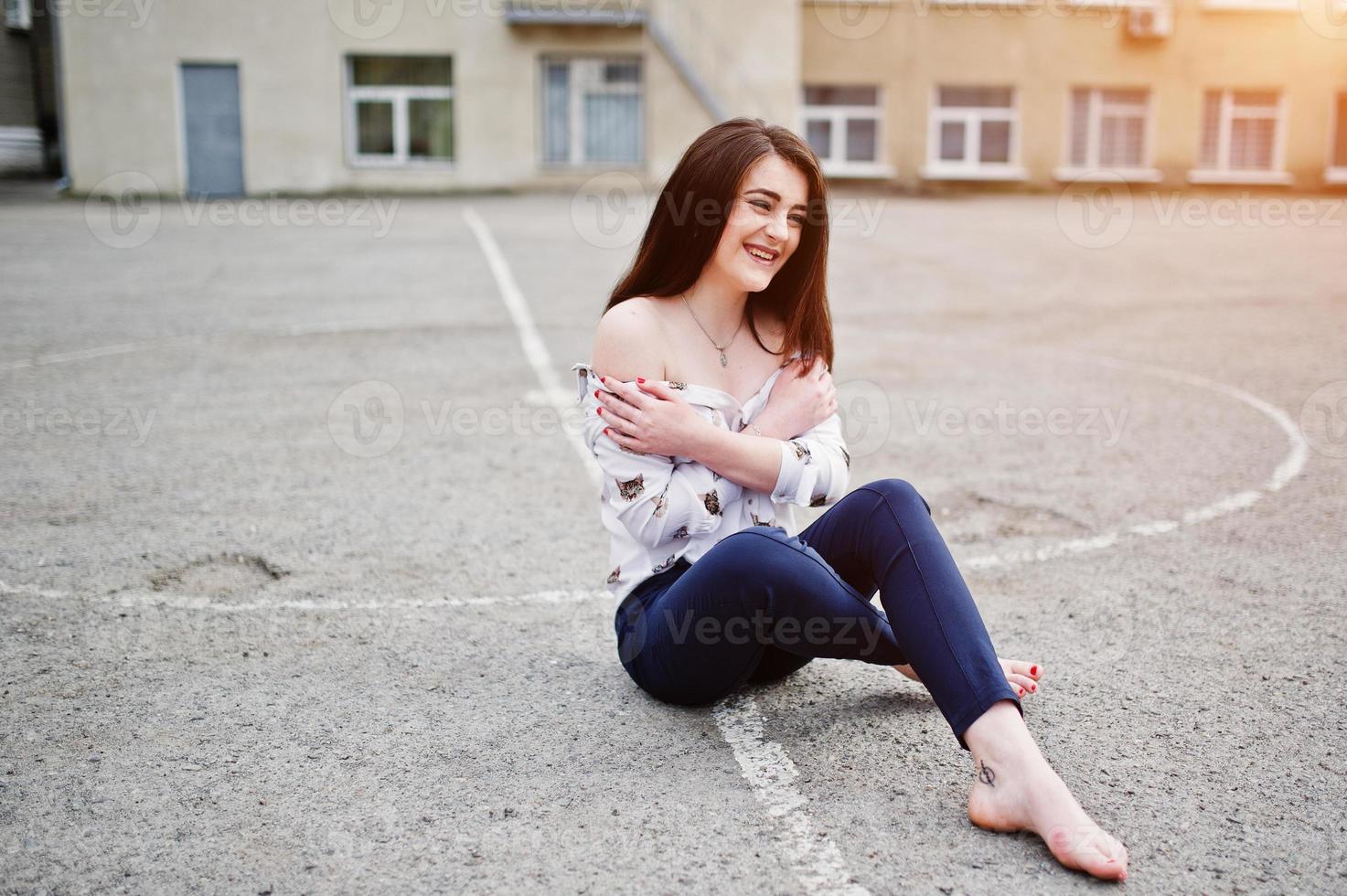 junge stilvolle Teenager-Brünette auf Hemd, Hose und Schuhen mit hohen Absätzen, sitzend auf Bürgersteig und gestelltem Hinterhof der Hintergrundschule. Straßenmode-Modellkonzept. foto