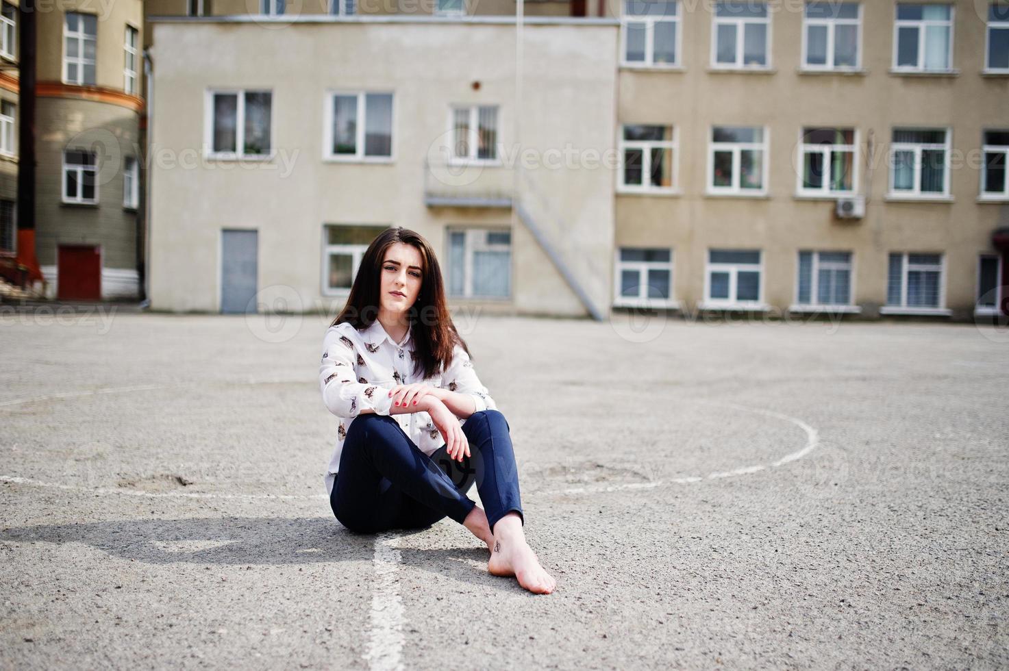 junge stilvolle Teenager-Brünette auf Hemd, Hose und Schuhen mit hohen Absätzen, sitzend auf Bürgersteig und gestelltem Hinterhof der Hintergrundschule. Straßenmode-Modellkonzept. foto