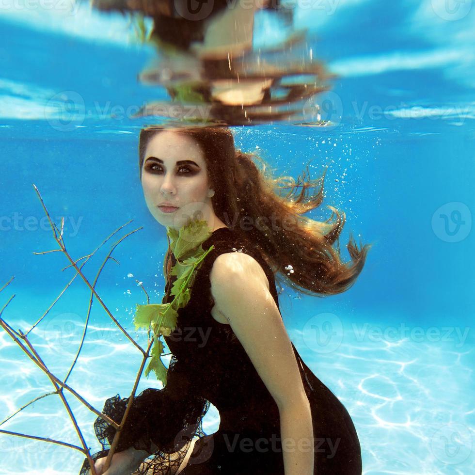 Unterwassermodeporträt einer schönen blonden jungen Frau in schwarzem Kleid mit Weinblättern foto