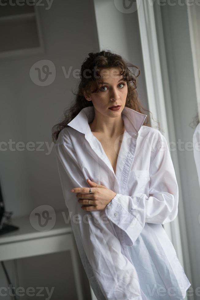 Attraktive sexy Brünette in weißem Hemd, die provokant posiert, in der Nähe eines Fensters, Studioaufnahme. Porträt einer sinnlichen Frau mit langen Haaren, in klassischer Boudoir-Szene, schaute aus dem Fenster foto