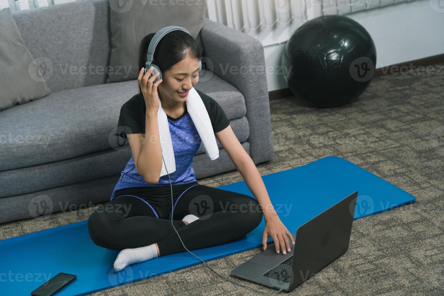 asiatische schöne junge frau, die laptop und kopfhörer verwendet, hört musik, während sie zu hause übung yoga praktiziert. foto